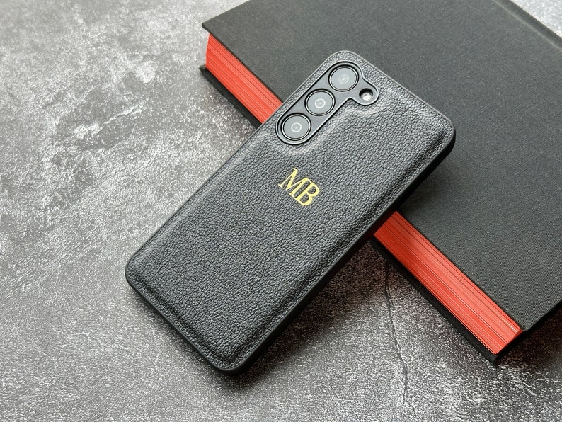 May include: A black leather phone case with a gold monogram "MB" and a camera lens cutout.