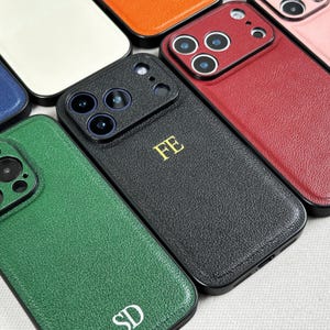 May include: A selection of leather phone cases in colours such as black, green, red, and orange. The black case displays the gold initials "FE", and the green case has "SD". The cases are designed for a smartphone with a camera bump.