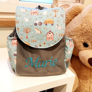 Könnte beinhalten: Ein hellblauer Rucksack mit einem Bauernhof-Motiv aus Traktoren, Scheunen und Tieren. Die Vorderseite ist grau und mit dem Namen Marie in leuchtend blauem Schreibschrift-Stick versehen. Ein kleines Etikett zeigt den Schriftzug Handmade unten.