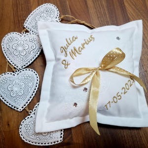 May include: White pillow with gold embroidery that reads "Julia & Marius 17-05-2023". The pillow is decorated with a gold ribbon and rhinestones. The pillow is surrounded by white metal hearts.