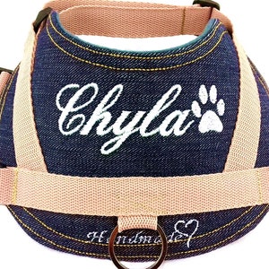 Può includere: Un'imbracatura per cani in denim blu con un'impronta di zampa ricamata in bianco e il nome "Chyla". L'imbracatura ha una cinghia regolabile rosa chiaro e un anello a D argentato.