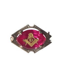 May include: A vintage brooch with a silver-toned frame and a vibrant pink center. The center features a gold-colored Masonic symbol. The brooch has a decorative, geometric design around the pink center.
