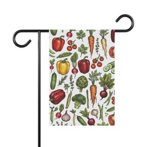 Op de afbeelding: Een rechthoekige tuinvlag hangend aan een zwarte metalen standaard, met een herhalend patroon van kleurrijke handgetekende groenten, waaronder rode en gele paprika's, wortels, radijsjes, broccoli, artisjokken, komkommers en diverse kruiden.