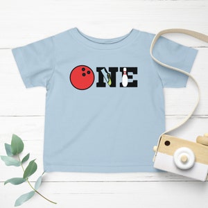 May include: Light blue short-sleeved t-shirt with the word "ONE" spelled out in black letters. The "O" is a red bowling ball, the "N" is a bowling shoe, and the "E" is a bowling pin.
