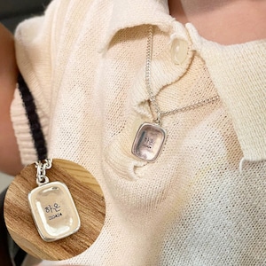 May include: Silver necklace with a rectangular pendant that says "하은" in Korean script. The pendant is on a silver chain and is worn by a person.
