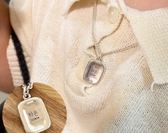 May include: Silver necklace with a rectangular pendant that says "하은" in Korean script. The pendant is on a silver chain and is worn by a person.