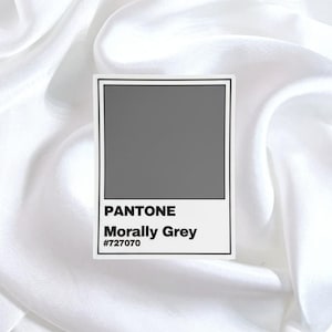 May include: A sticker featuring a Pantone color swatch in Morally Grey (#727070). The design includes the Pantone logo and color name in black text on a white background. The sticker is set against a white, silky fabric.