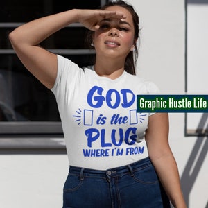 May include: White t-shirt with blue text that reads "GOD is the PLUG WHERE I'M FROM". The text is in a bold, blocky font. The words "Graphic Hustle Life" are in smaller text in the upper right corner of the shirt.
