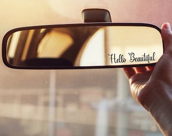 May include: A black car rearview mirror with the words "Hello Beautiful" in a cursive font. The mirror is held by a hand, with the car's interior and exterior visible in the reflection.