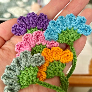 May include: Five crocheted flower embellishments in various colors, including purple, blue, pink, green, and orange. The flowers are connected by a green crocheted stem.