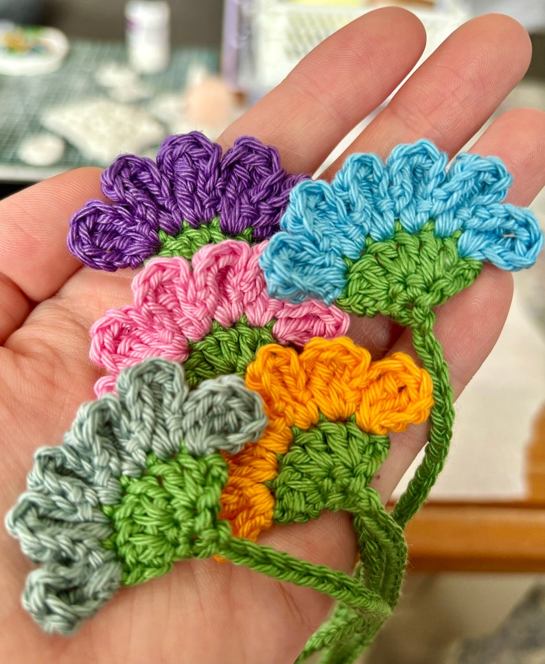 May include: Five crocheted flower embellishments in various colors, including purple, blue, pink, green, and orange. The flowers are connected by a green crocheted stem.