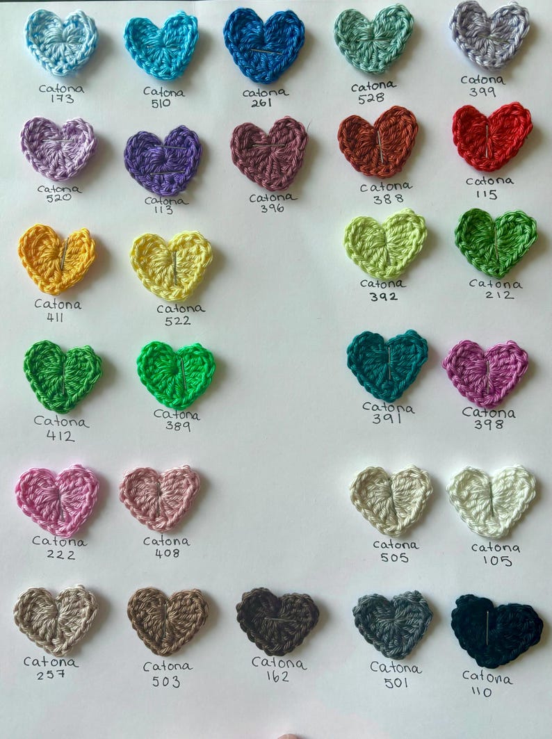 May include: A collection of crocheted hearts in various colors, each labeled with the corresponding Catona yarn number.