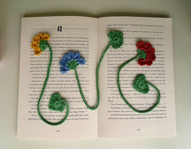 May include: A green, red, and blue crocheted bookmark with flower designs. The bookmark is hanging from a book with the text "48" and "172" visible.