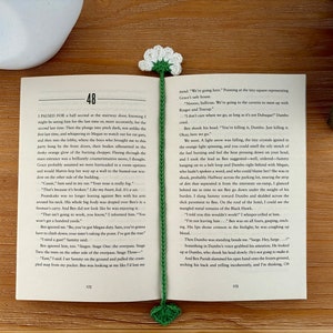 May include: A close-up of an open book with a green crocheted bookmark. The bookmark has a white flower on top. The book is open to page 112 and the text on the page reads "I paused for a half second on the stairway down, knowing I might be using him for the last time or, more accurately, for the second last time. Then the plunge into pitch dark, no under the first time, and whispering to Megan to watch out for gas, and then into the lobby, where the boys who brought me to the party hung by the front doors, their bodies silhouetted in the dusky orange glow of the burning chopper. Fleeting through the main entrance was a brilliantly communicative move, I thought. Grace probably assumed we were barricaded in a room, upstairs and would Marsis-hop her way up a wall to the busted-out window on the other side of the building."