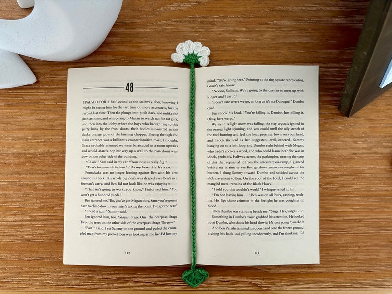 May include: A close-up of an open book with a green crocheted bookmark. The bookmark has a white flower on top. The book is open to page 112 and the text on the page reads "I paused for a half second on the stairway down, knowing I might be using him for the last time or, more accurately, for the second last time. Then the plunge into pitch dark, no under the first time, and whispering to Megan to watch out for gas, and then into the lobby, where the boys who brought me to the party hung by the front doors, their bodies silhouetted in the dusky orange glow of the burning chopper. Fleeting through the main entrance was a brilliantly communicative move, I thought. Grace probably assumed we were barricaded in a room, upstairs and would Marsis-hop her way up a wall to the busted-out window on the other side of the building."