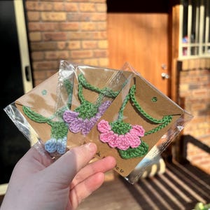 May include: Three crocheted flower necklaces in pink, purple, and blue, displayed on brown card stock with a white background. The necklaces are packaged in clear plastic bags.