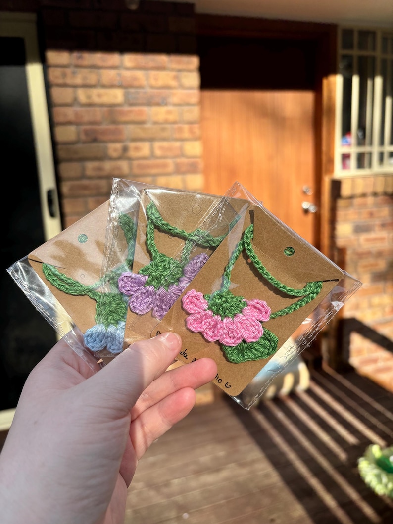 May include: Three crocheted flower necklaces in pink, purple, and blue, displayed on brown card stock with a white background. The necklaces are packaged in clear plastic bags.