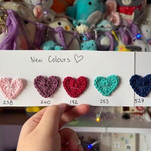 May include: A white card with the text "New Colours" and a heart symbol. Five crocheted hearts in different colors are displayed on the card with their corresponding numbers: 238, 240, 192, 253, and 527.