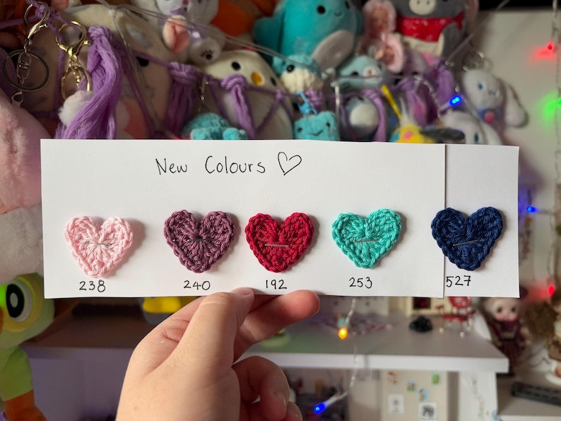 May include: A white card with the text "New Colours" and a heart symbol. Five crocheted hearts in different colors are displayed on the card with their corresponding numbers: 238, 240, 192, 253, and 527.