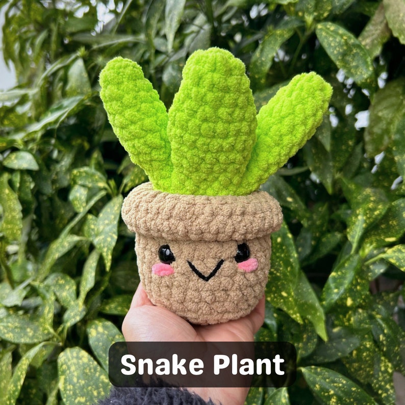 May include: A crocheted snake plant with a cute face. The plant is green and brown and has a happy expression.