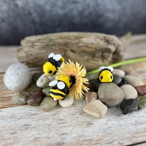 Op de afbeelding: Drie kleine, handgemaakte hommels van polymer clay. De bijen zijn geel en zwart met witte accenten. Ze zijn gerangschikt op een houten oppervlak met een bloem en stenen.