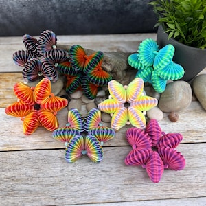 Può includere: Una collezione di decorazioni a farfalla di carta colorate in varie tonalità di rosso, arancione, verde, blu, viola e giallo. Le farfalle sono disposte su una superficie di legno, con una piccola pianta in vaso sullo sfondo.