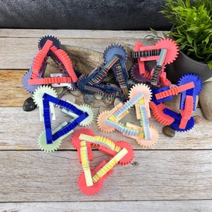 Può includere: Un set di otto giocattoli a forma di ingranaggio triangolare colorati. Ogni ingranaggio è realizzato in plastica e ha un colore e un motivo diversi. Gli ingranaggi sono disposti in cerchio su una superficie di legno.