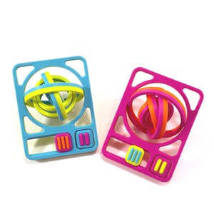 Puede incluir: Dos juguetes fidget rectangulares con esquinas redondeadas. El juguete de la izquierda es azul claro con un anillo giratorio amarillo y azul en el centro y dos botones pequeños. El de la derecha es magenta con un anillo giratorio naranja y magenta y dos botones.