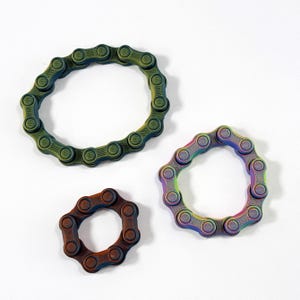 Puede incluir: Tres juguetes fidget circulares construidos con eslabones met&aacute;licos interconectados. Un anillo grande presenta un acabado verde mate, un anillo mediano muestra un degradado arco&iacute;ris vibrante y un anillo peque&ntilde;o tiene un acabado cobrizo oscuro texturizado.