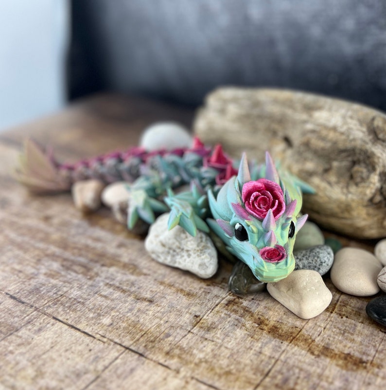 May include: A small, green and pink dragon figurine with a rose on its head. The dragon is lying on a wooden surface with small rocks around it.