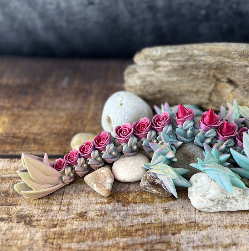 May include: A pink and green succulent floral arrangement with a leafy tail. The arrangement is made of polymer clay and is displayed on a wooden surface with rocks.