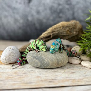 Puede incluir: Dos llaveros de camaleón coloridos, uno verde y otro azul, con detalles intrincados. Los llaveros están sobre una piedra gris con un fondo de madera.