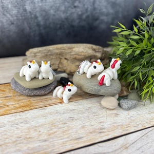 Può includere: Quattro figurine di unicorno bianche con corna dorate e accenti rossi. Gli unicorni sono seduti su rocce grigie.