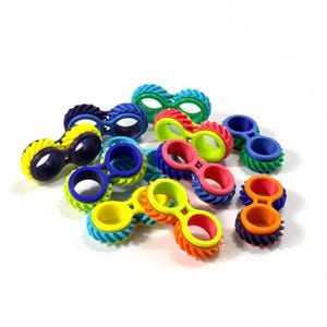 Könnte beinhalten: Eine Sammlung von mehrfarbigen Fidget-Spielzeugen, die jeweils aus zwei verbundenen Ringen mit einem texturierten, zahnradartigen Außenrand bestehen. Die Spielzeuge sind in verschiedenen leuchtenden Farbkombinationen wie Blau, Grün, Gelb, Lila, Pink und Orange gehalten.