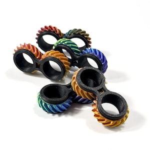 Puede incluir: Un conjunto de tres juguetes fidget de doble anillo, cada uno con dos aberturas circulares negras conectadas por un puente central. Los bordes exteriores de los anillos tienen una textura en espiral similar a engranajes con colores degradados.