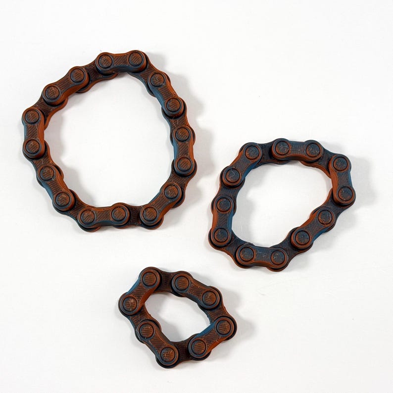 Puede incluir: Tres bucles circulares construidos con eslabones de cadena de bicicleta de color marr&oacute;n oscuro y negro. Los eslabones est&aacute;n dispuestos en tama&ntilde;os variables, con un anillo grande, uno mediano y uno peque&ntilde;o, cada uno con un aspecto met&aacute;lico.