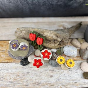 Può includere: Assortimento di elastici per capelli a forma di fiore colorati. Gli elastici per capelli sono realizzati in pasta polimerica e presentano una varietà di disegni di fiori, tra cui tulipani, girasoli e margherite. Gli elastici per capelli sono esposti su una superficie di legno con piccole pietre.