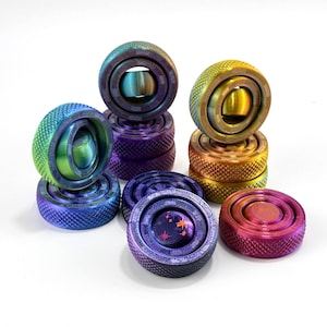 Puede incluir: Una colección de fidget spinners circulares de metal con acabados iridiscentes tipo arcoíris. Cada spinner tiene un borde exterior texturizado y moleteado con un centro giratorio suave. Los colores van desde azules y morados profundos hasta verdes y dorados vibrantes.