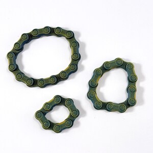 Puede incluir: Tres bucles circulares construidos con eslabones de cadena de bicicleta interconectados en color verde oscuro y amarillo. Los eslabones presentan una superficie texturizada y estriada, dispuestos en tres tama&ntilde;os distintos formando anillos flexibles.