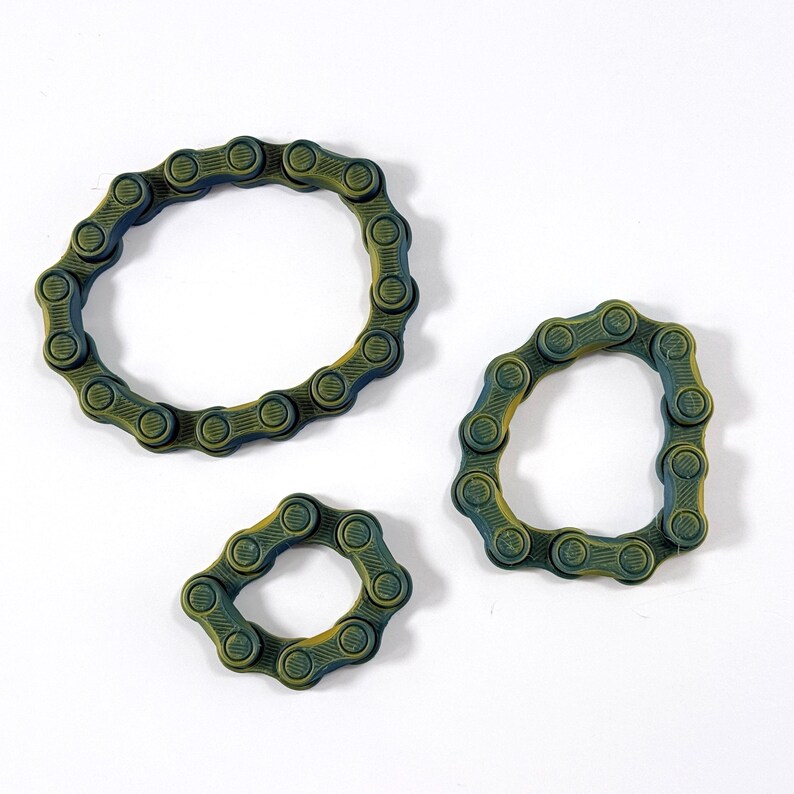 Puede incluir: Tres bucles circulares construidos con eslabones de cadena de bicicleta interconectados en color verde oscuro y amarillo. Los eslabones presentan una superficie texturizada y estriada, dispuestos en tres tama&ntilde;os distintos formando anillos flexibles.