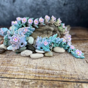May include: Two small, blue and green, flower-adorned, dragon figurines with pink flowers. The dragons are sitting on a wooden surface with small stones.