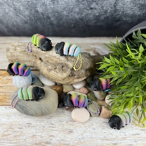 Può includere: Una collezione di sette portachiavi a forma di millepiedi, di colore nero iridescente e arcobaleno. I portachiavi sono realizzati in pasta polimerica e misurano circa 5 cm di lunghezza. Sono esposti su una superficie di legno con piccole pietre.
