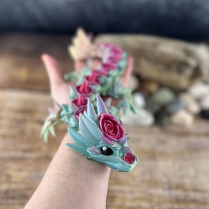 May include: A hand holding a small, green and pink dragon figurine with rose decorations. The dragon has a cute face with black eyes and a pink nose.