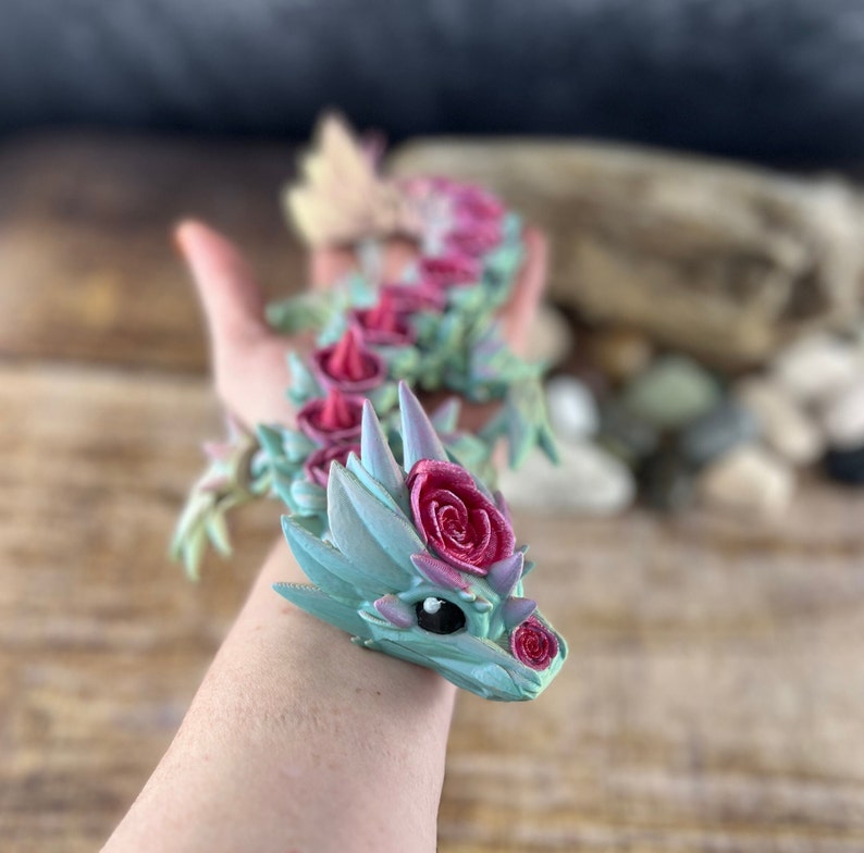 May include: A hand holding a small, green and pink dragon figurine with rose decorations. The dragon has a cute face with black eyes and a pink nose.