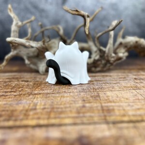 May include: A white ghost figurine with a black cat tail peeking out from under the sheet. The ghost is sitting on a wooden surface.