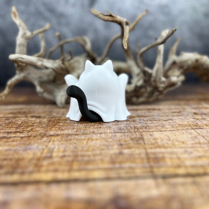 May include: A white ghost figurine with a black cat tail peeking out from under the sheet. The ghost is sitting on a wooden surface.