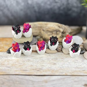 Può includere: Otto figurine di drago in miniatura, ciascuna in un contenitore bianco a forma di uovo. I draghi sono neri e rosa con squame iridescenti. Le uova sono disposte in fila su una superficie di legno.