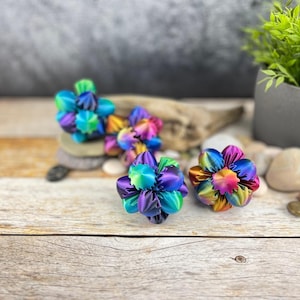 Può includere: Tre palline di carta piegate a forma di fiore, iridescenti e multicolori. Le palline sono fatte di carta piegata e hanno una finitura lucida e metallica. Le palline sono disposte in fila su una superficie di legno.