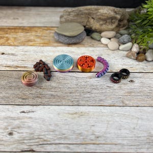 Può includere: Una collezione di giocattoli sensoriali colorati, tra cui uno spinner a vortice arcobaleno, uno spinner a forma di stella rosso e arancione, una molla viola e nera e due anelli neri e argentati.