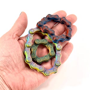 Puede incluir: Tres juguetes antiestr&eacute;s circulares construidos con eslabones de cadena de bicicleta de metal interconectados. Un anillo presenta un degradado azul y cobre, otro es verde oscuro s&oacute;lido y el tercero muestra un acabado arco&iacute;ris.